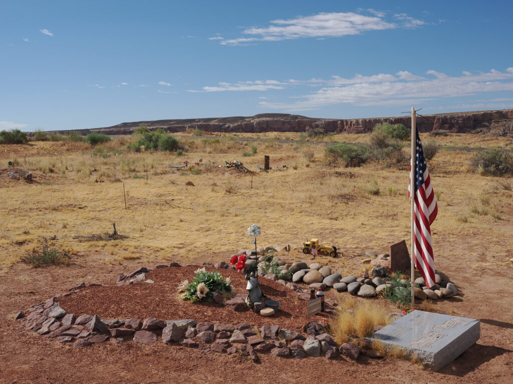 Photo taken facing south with a Bluff Cemetery plot featuring heart-shaped rock memorial, decorative memorial items, an an American flag with red rock cliffs in the background.