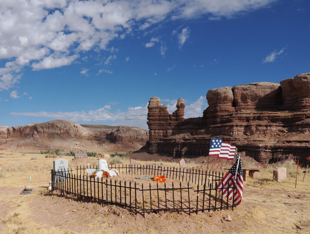 Photo taken facing northwest of a Bluff Cemetery plot with a black picket fence, two American Flags, headstones, and the large "Locomotive Rock" and Bluff cliffs in the background.
