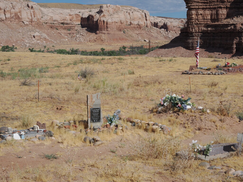 Photo taken facing north with multiple headstones and memorial items pictured, as well as natural desert landscape with dry grasses and the Bluff cliffs in the background.